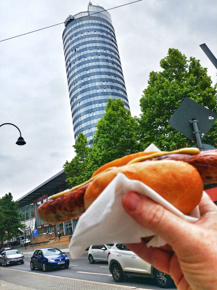 Männerhand mit Roster (Thüringische Rostbratwurst) vor dem JenTower (Keksrolle), einem der Wahrzeichen Jenas