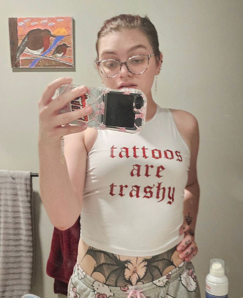 Alternative Femme with tattoos on stomach and arm poses in mirror wearing shirt that reads "Tattoos are Trashy"