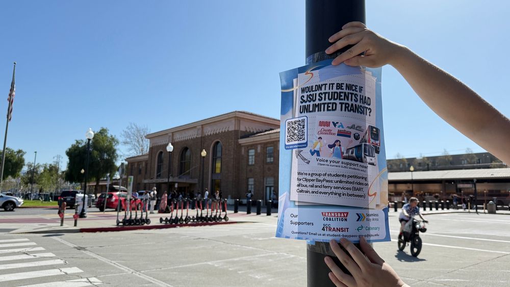 Poster being hung up with a call-to-action saying “Wouldn’t it be nice if SJSU students had unlimited transit?” outside the historic San Jose Diridon station.
