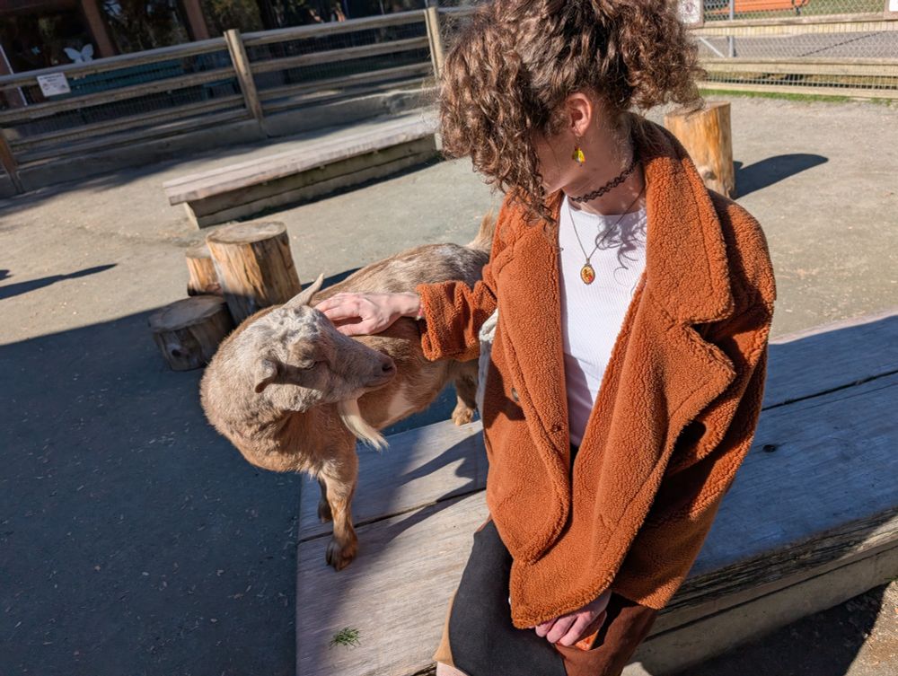 fern sitting on a bench petting a goat who is standing next to her and they are looking at each other