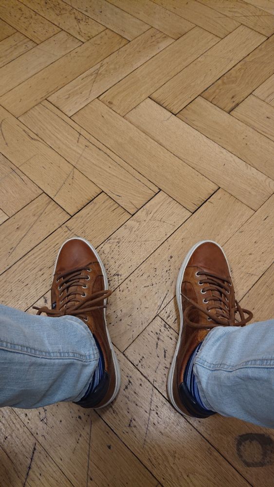 Blick von oben auf meine Unterschenkel in hellblauen Jeans und die Füße in braunen Sneakers auf Fischgrätparkett. 