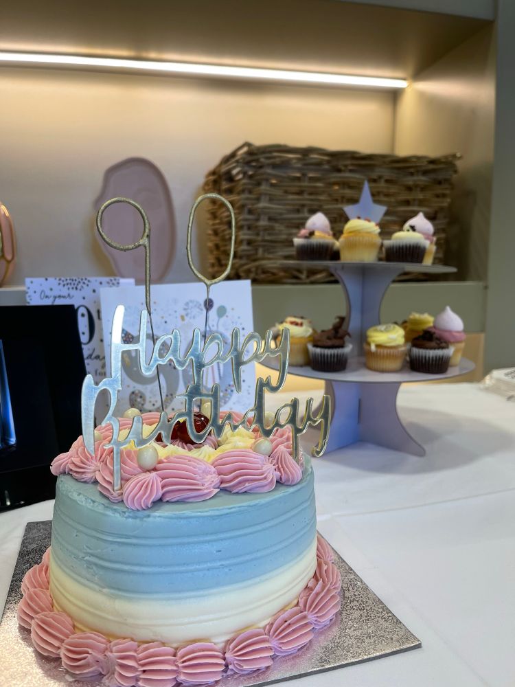 A round cake with multi-coloured icing with a Happy Birthday banner. Behind a cake stand with cupcakes.