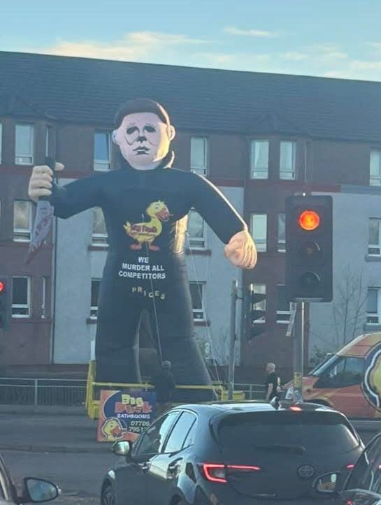 A huge blow up Michael Myers in the street, wielding a knife, with a duck logo on his shirt. Underneath that, white text reads 'we murder all competitors.... Prices.'