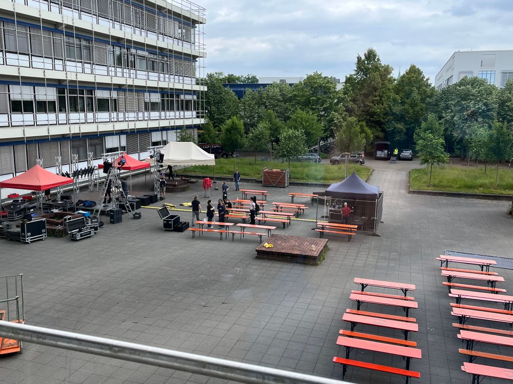 Blick in den Innenhof des Informatikgebäudes. Man sieht links eine Bühne mit Zeltdächern, in der Mitte ein Pavillon für die Soundtechnik und rechts viele Bierbänke. Dazwischen laufen Menschen, die den Aufbau bewerkstelligen.