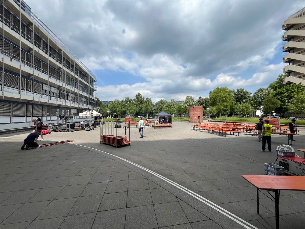 Blick in den Innenhof des Informatikgebäudes. Man sieht links eine Bühne mit Zeltdächern, in der Mitte ein Pavillon für die Soundtechnik und rechts viele Bierbänke. Dazwischen laufen Menschen, die den Aufbau bewerkstelligen.