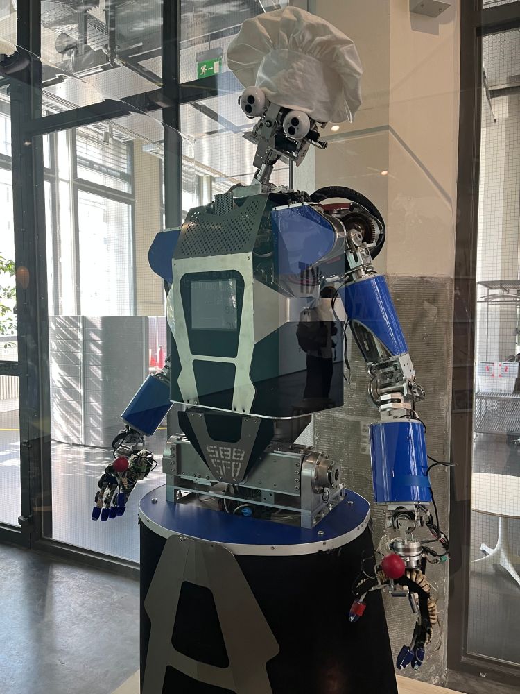 Der humanoide Roboter ARMAR -III steht in einer hohen Vitrine. Er hat eine Kochmütze auf. Deutlich zu erkennen seine Arme und Hände sowie seine fahrbare Plattform.