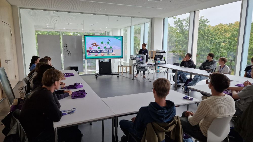 In einem Seminarraum sitzen einige Personen an in U-Form angeordneten Tischen. Eine Dozenten präsentiert eine Folie auf einem großen Monitor.