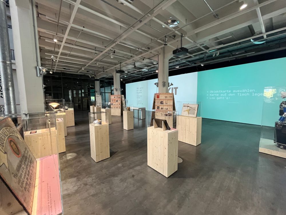 Ausstellungsfläche "200 Jahre KIT | 100 Objekte. Teile des Ganzen" im ZKM Karlsruhe. Man sieht Podeste mit Exponaten und im Hintergrund große Projektionsflächen.