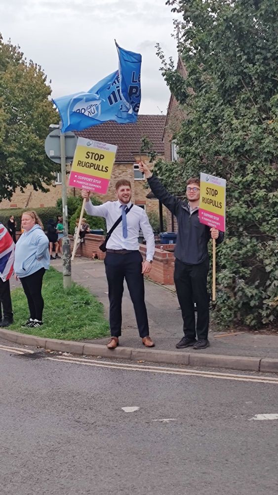 Two young men, one wearing a tie, holding fake stand up to racism placards, reading 'Stop rugpulls. Support STUF. Pumpfun supports migrants'. Behind a Reform party banner waves.