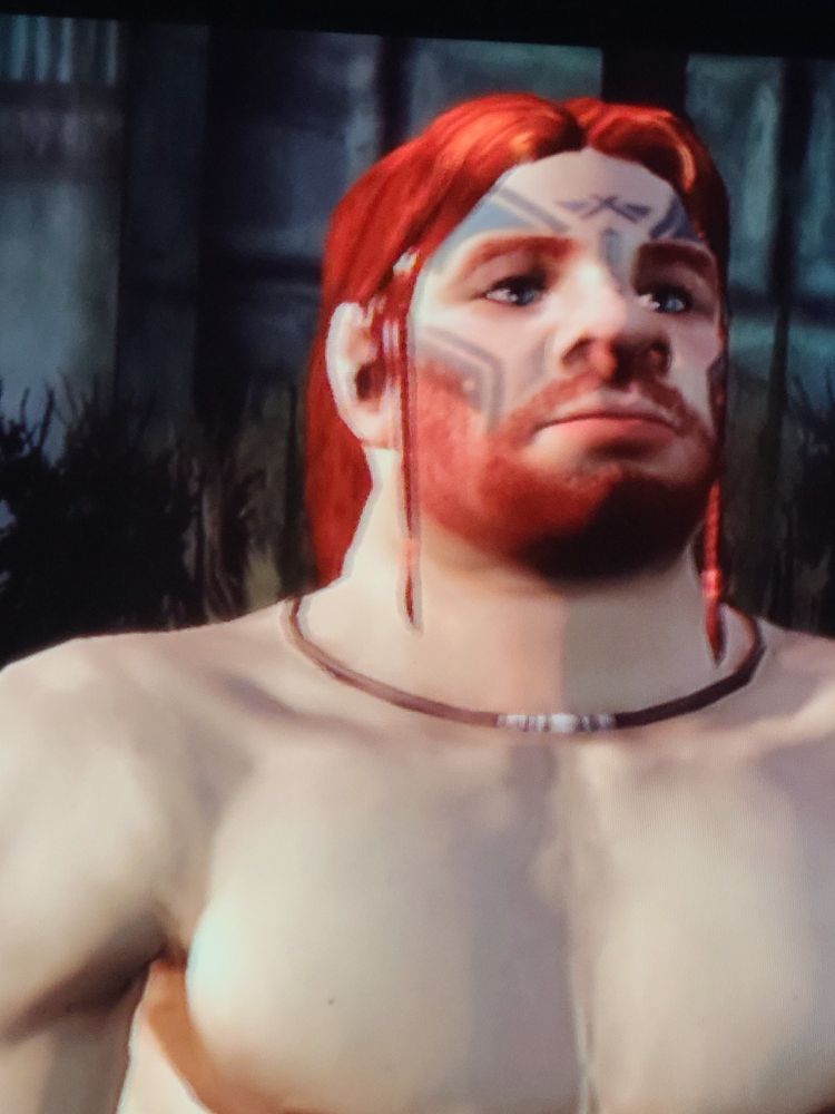 A closeup on the face and chest of a red-haired dwarf with green eyes and large blue facial tattoos around his eyes and on his nose. The dwarf is shirtless.