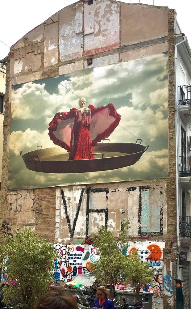 auf einer hohen feuermauer ist am oberen drittel ein kunstwerk angebracht. es zeigt eine opernsängerin im roten langen gewand mit erhobenen armen im ausgebreiteten übermantel. die figur steht in einer landestypischen paella-pfanne.