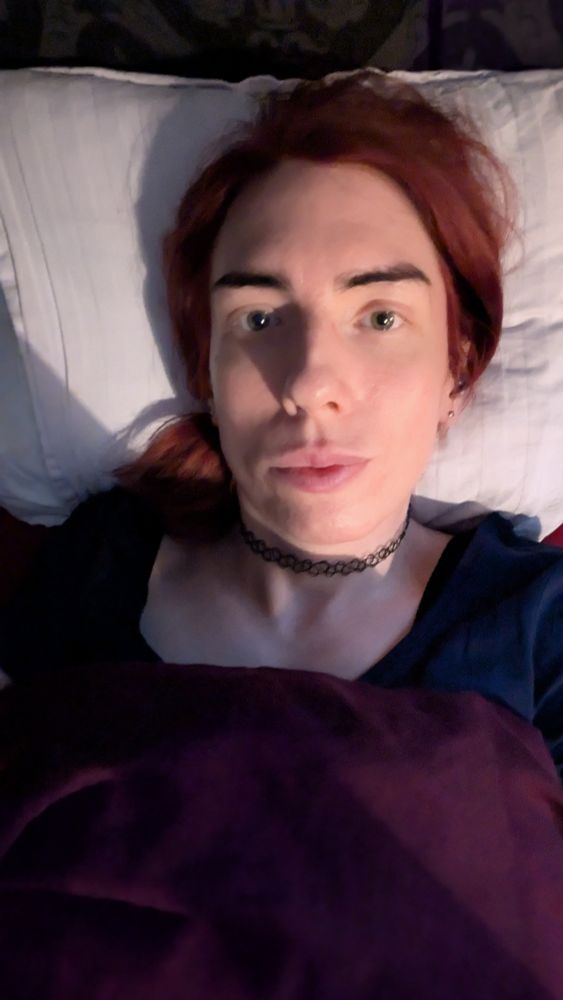 A woman in bed for a nap. She is wearing a blue top and a choker. Her hair is red.