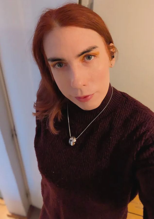 A red haired woman in a burgundy sweater dress. She has a silver necklace. She has pink red lips and red-orange eyeshadow.