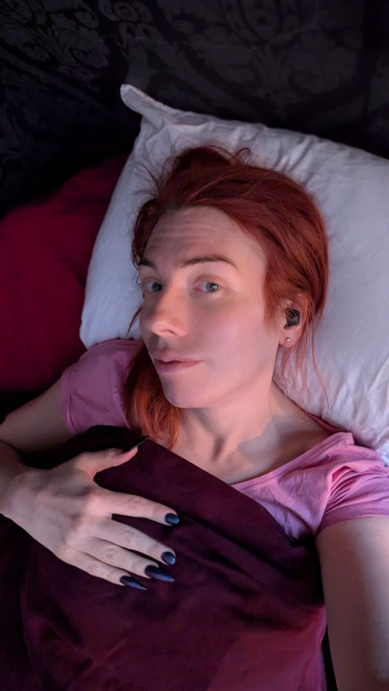 A red haired woman with a pink t-shirt lying in a bed with the duvet pulled up to her chest. 