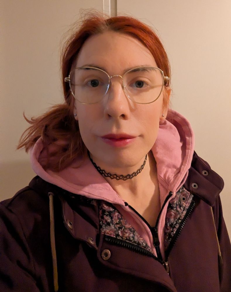 A red haired woman with glasses in a burgundy jacket over a pink hoodie. She has a TheTransChoker™ on.