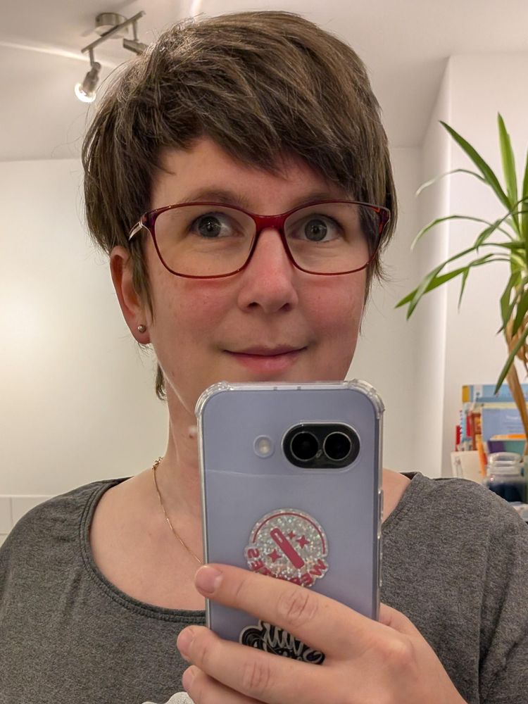 Sara taking a mirror selfie in her bathroom wearing a pair of deep red thin framed oval-rectangle glasses.