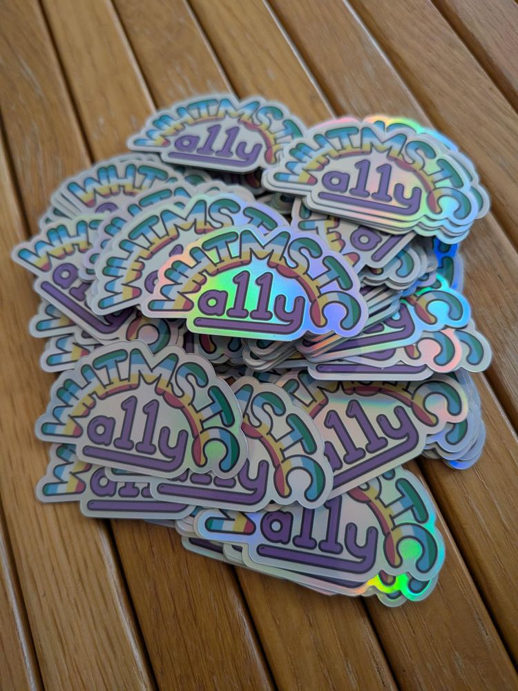 A pile of whimsica11y stickers, their holographic effect showing a gentle rainbow across several of them.

On each sticker the letters WHIMSIC form an arch, within which are the disability pride colours green, blue, white, yellow and red, in rainbow-style stripes. Under the arch is "a11y" in the named colour Rebecca purple.