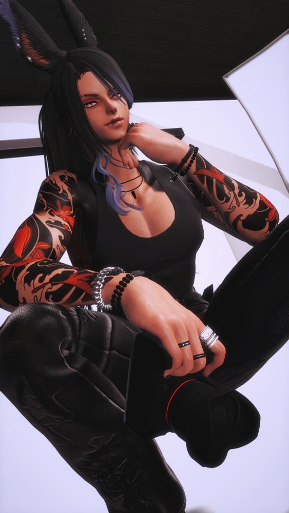 Cai sits confidently in a photography studio. His long dark hair, streaked with slate blue, frames a sharp gaze, while intricate red-and-black tattoos of water and maple leaves wind around his arms. Dressed in sleek black attire and adorned with layered bracelets rings, lip and ear piercings, he holds a camera in one hand, exuding a cool, artistic presence against the studio’s clean, modern backdrop.