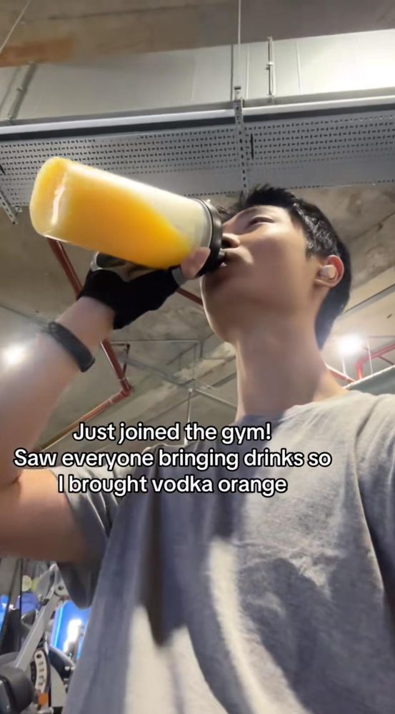 A person in a gym is drinking from a large bottle filled with an orange-colored beverage. They are wearing a gray shirt and gloves, and there is gym equipment in the background. Text on the image humorously mentions bringing vodka orange to the gym.