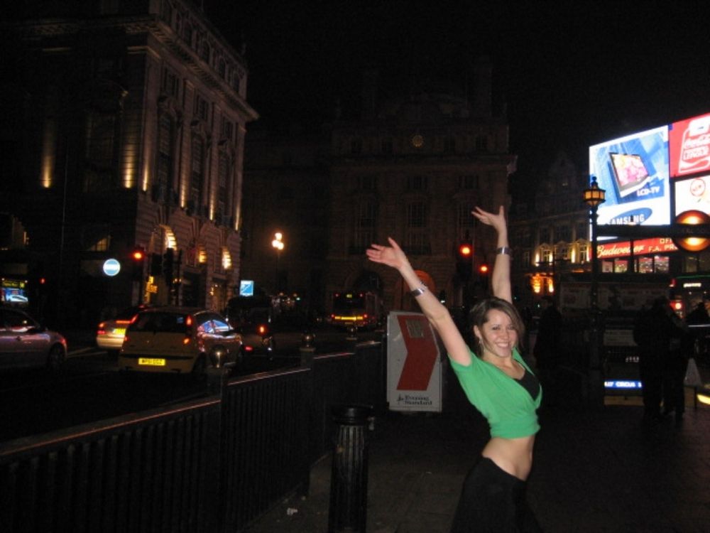 Cooper streaming wearing a midriff bearing outfit throwing both her hands in the air and smiling at the camera standing in Piccadilly circus, London