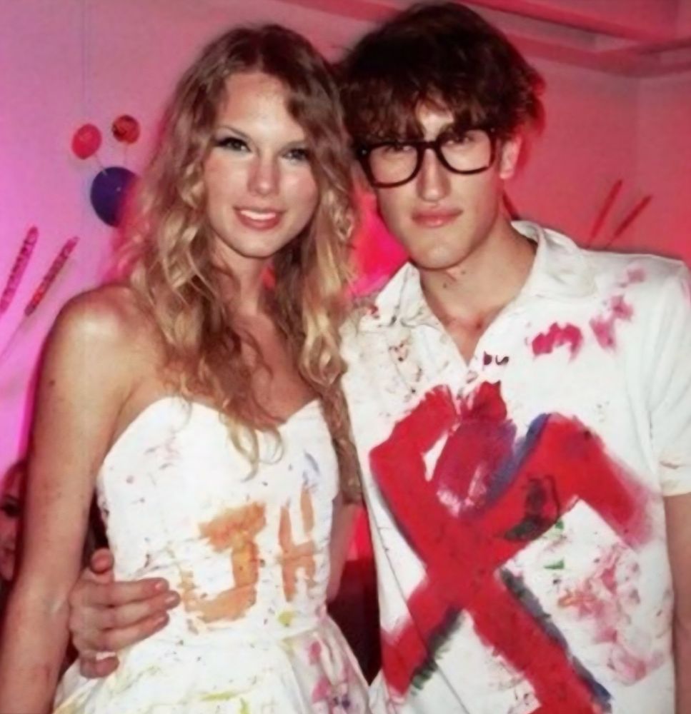 Taylor Swift and male model pictured posting for a photo each with an arm around each other during Katy Perry's 25th birthday party. There is a pink walled background decorated with candy, while in front, Taylor Where's a white dress and a model wears a white polo. Both are completely covered in spattered paint with Taylor's dress saying JH on the front and the male model supporting large glasses and a shirt with an extremely large swashtika that takes up the entire front surface of the shirt.