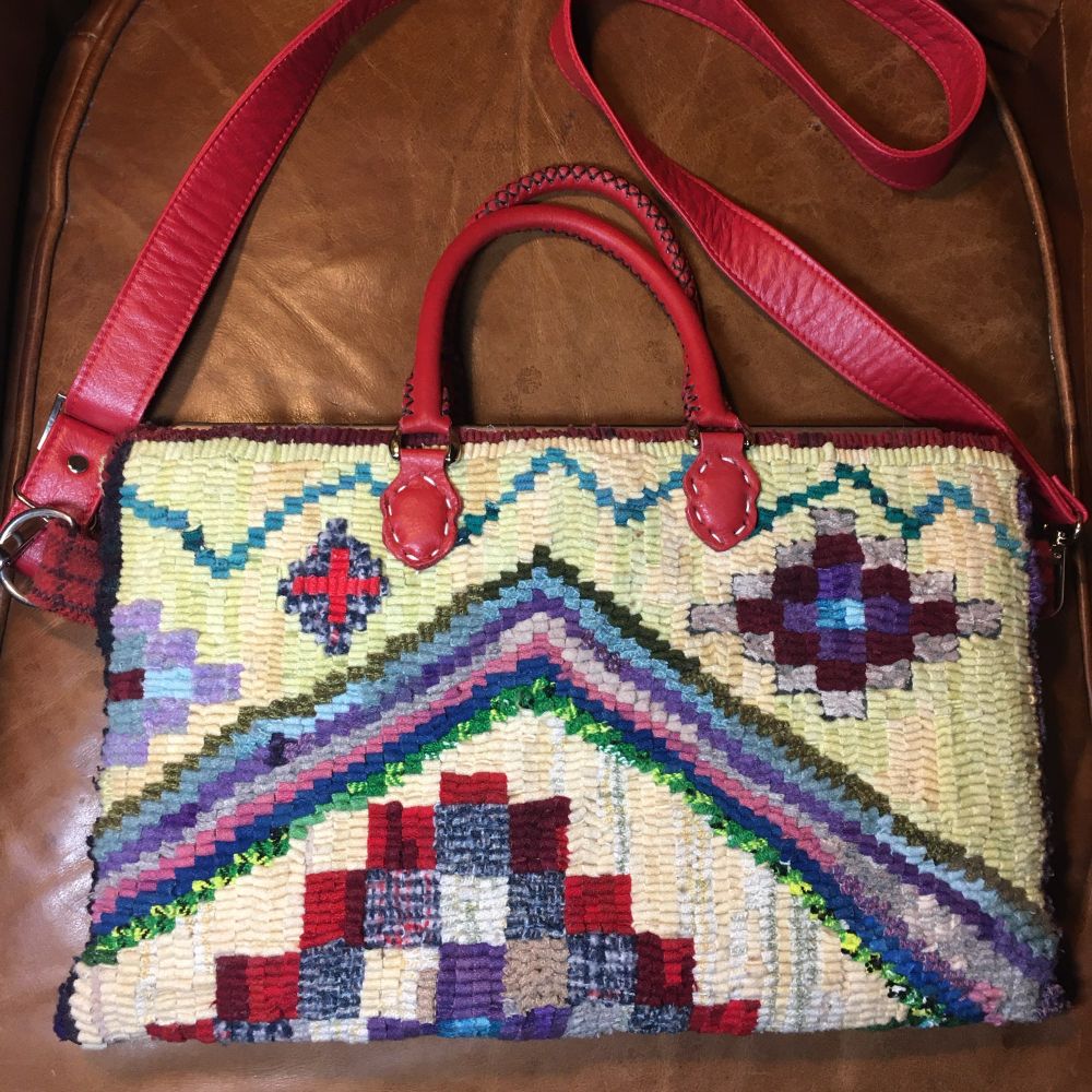 One side of a rectangular hooked carpetbag with crimson-red rolled-leather handles and long matching shoulder strap, laying flat on the seat of a butterscotch-leather chair. Design is described in 3rd photo’s alt text. Hooked primarily using leftover strips, with all loops oriented the same way to mic weaving, I learned SO much about colour (as well as the history of the motifs) in this workshop with Jen Manuell. 