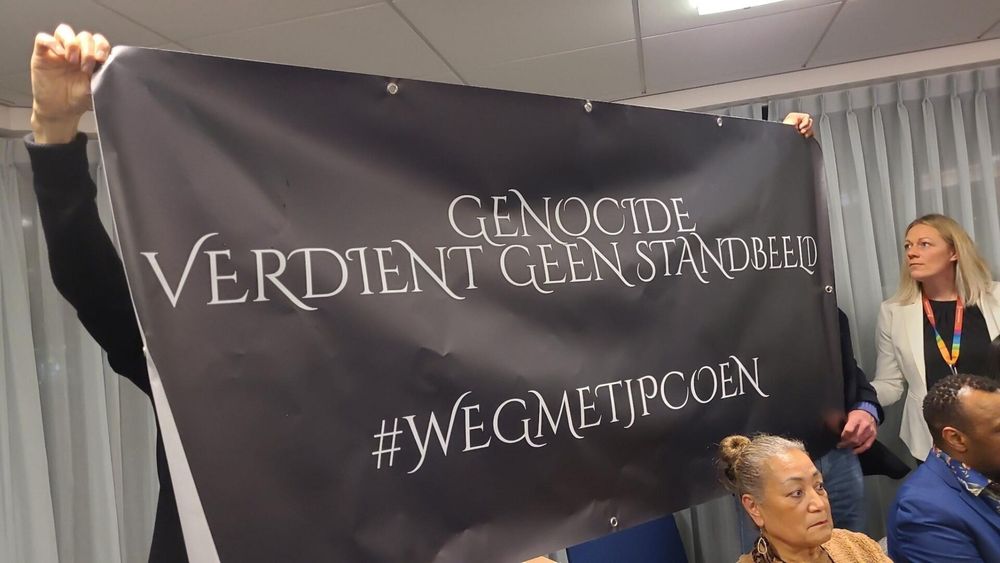 Zwart met wit beletterd spandoek in de raadszaal: 

            GENOCIDE 
VERDIENT GEEN STANDBEELD

#WEGMETJPCOEN