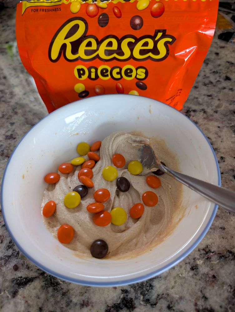 A bowl of vanilla oatmilk ice cream with peanut butter hand mixed together, and Reese's Pieces sprinkled on top. The bag of Reese's Pieces is behind the bowl