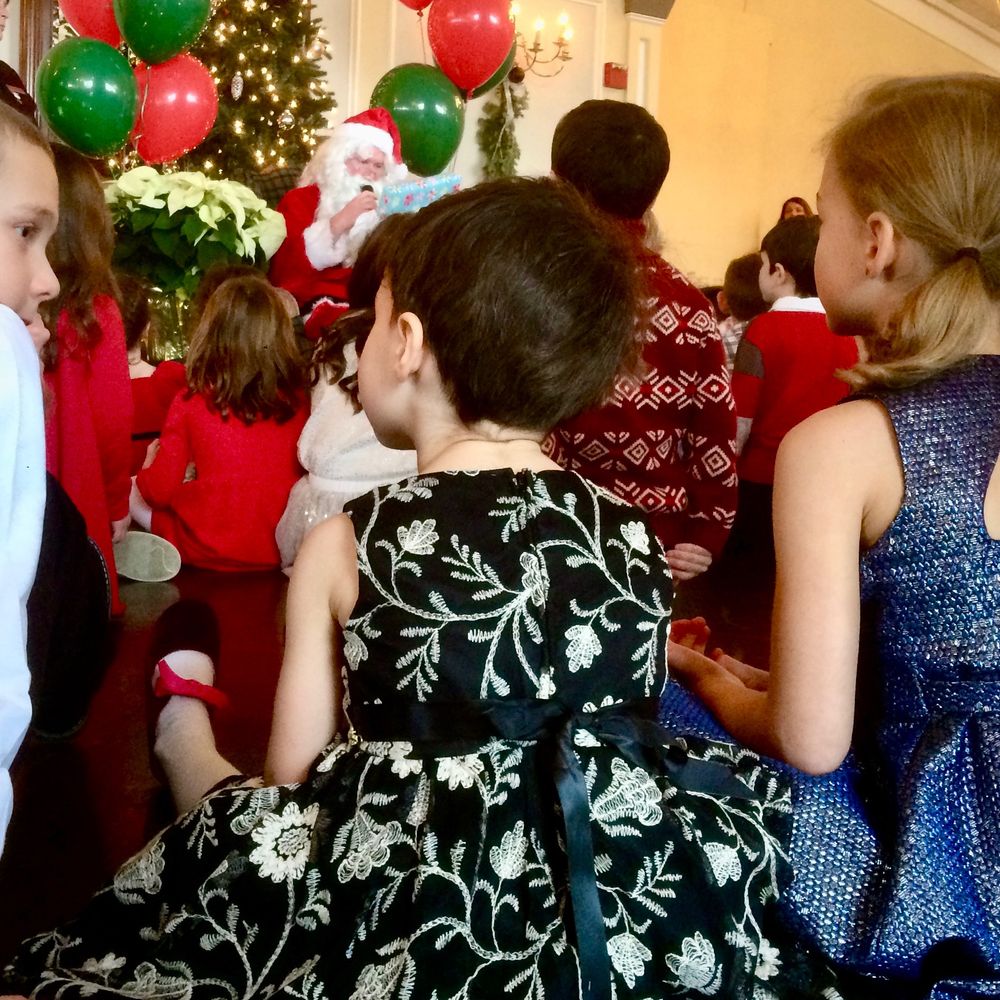 Picture of a kid from the back, wearing a holiday dress and a boys haircut. In the background is Santa handing out gifts.