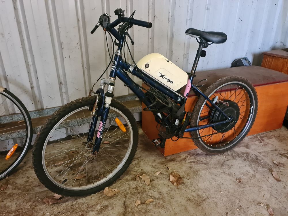 A low quality child-sized electric bike in a dirty garage. 24" tyres, K-mart mountain bike frame, with a lot of rust.