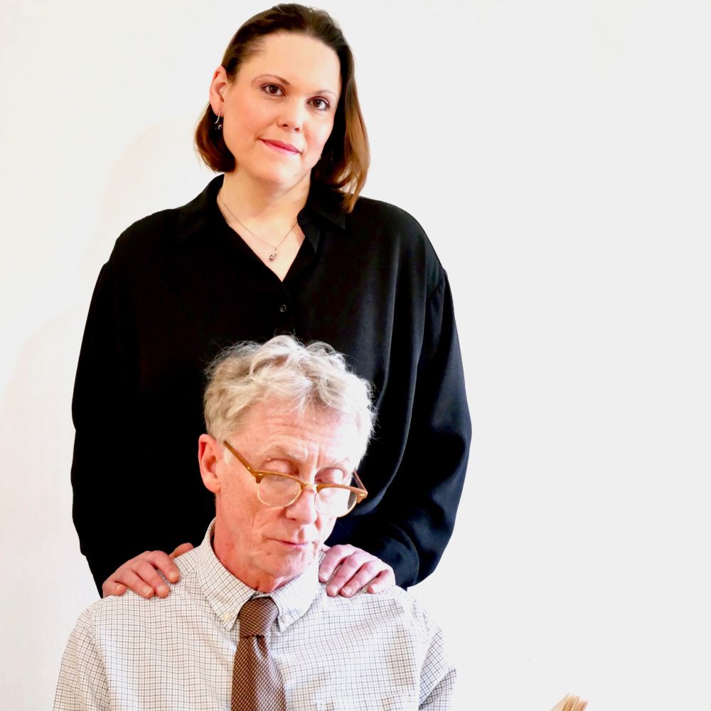 A man wearing glasses, and a shirt and tie is sitting down. A woman wearing a black shirt stands behind him with her hands on his shoulders. 