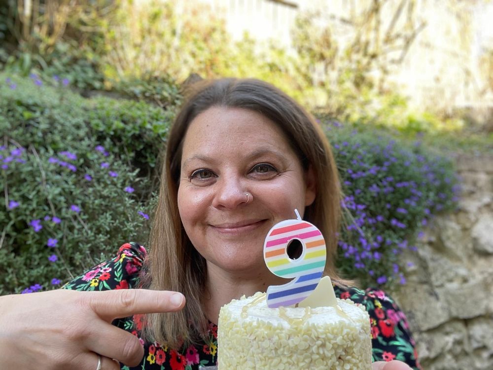 A photo of a woman standing outside and holding a cake. The cake has a number nine candle in it and the woman is pointing at it. She is wearing a floral dress and we can see a wall and shrubs in the background. 