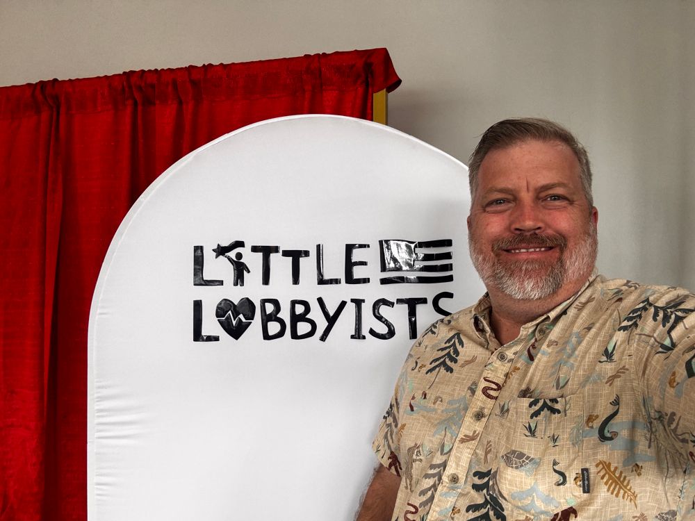 Older distinguished looking gentleman with a beard standing in front of a Little Lobbyists sign. 