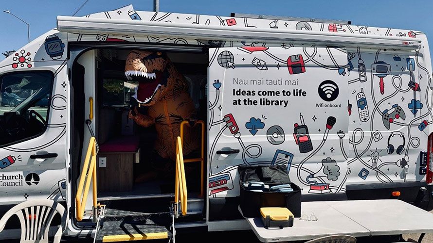 Photo of a library staff member in an inflatable dinosaur costume standing at the door way of the library tech van.