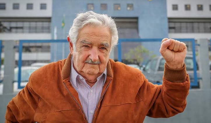 Pepe Mujica usa um suéter laranja e uma camisa de botão listrada por baixo. Ele está com o punho de seu lado esquerdo cerrado para cima
