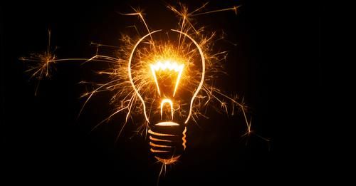 A clear electric light bulb with gold sparks jumping off of it.
