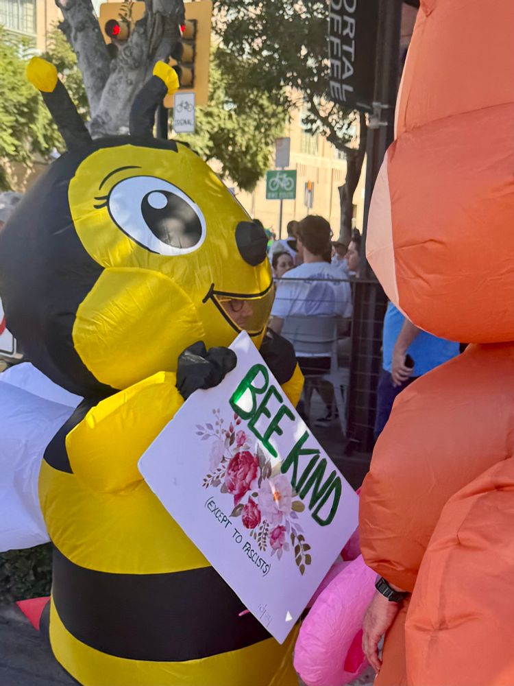 Protestor in a Bee costume holding a Bee Kind (except to racists) sign