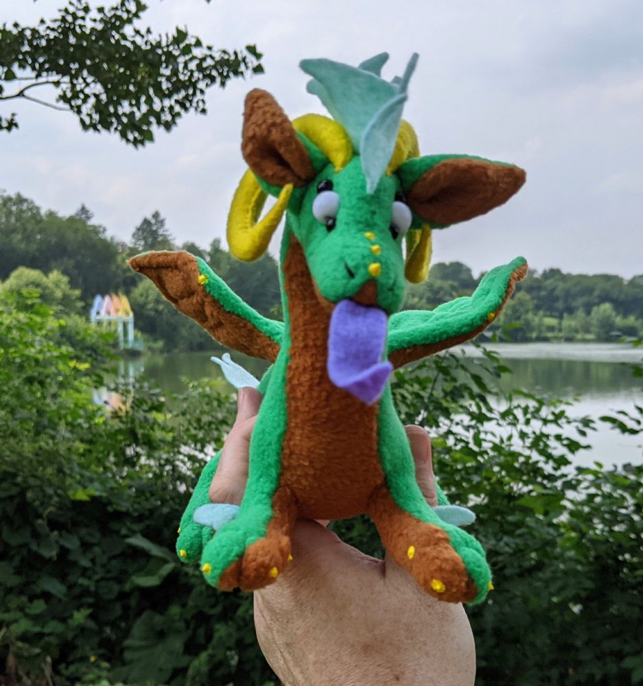 a green dragon plushie with a warm brown underbelly/underwings/inner ears, yellow horns/claws/markings, a purple tongue, aqua mane/tail/elbow tufts, and white eyes