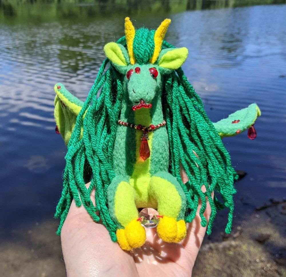 a green dragon-horse plushie holding a cut glass crystal between their cloven yellow hooves with a lake in the background. they are almost a bottle green, with a lighter yellowy green underbelly, red eyes/bead details/embroidery, and a necklace of red and gold beads