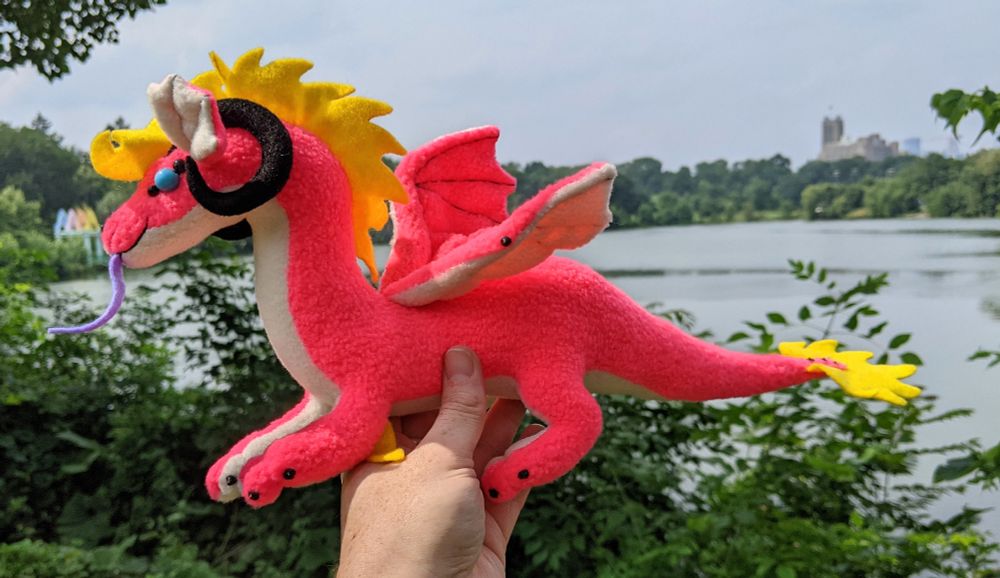 a bright neon pink dragon with a white underbelly/inner ears/underwings, purple tongue, yellow mane/tail/elbow tufts, black horns/claws/markings, sky blue eyes