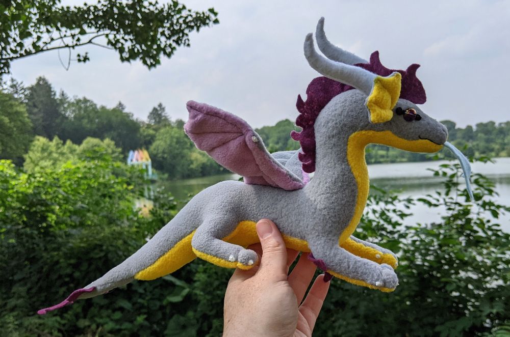 a soft gray dragon plushie with a honey yellow underbelly and inner ears, light purple/lilac underwings, raspberry colored mane/tailtuft/elbow tufts, white horns/claws/markings, and golden purple glass eyes