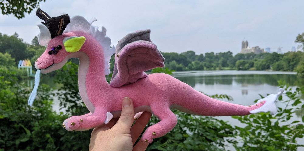 a pink dragon with a pastel stripes underbelly, aqua and yellow ears, gray and purple wings, white mane/tail/elbow tufts, gold claws/markings, and black & gold mothlike antennae and golden purple glass eyes