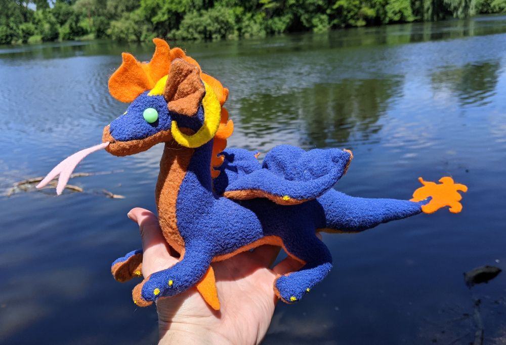 a deep blue dragon from the side, with a rusty orange underbelly/underwings/inner ears, a pink tongue, yellow horns/claws/details, a bright orange crest and tail tuft, orange elbow claws, and bright light aqua eyes