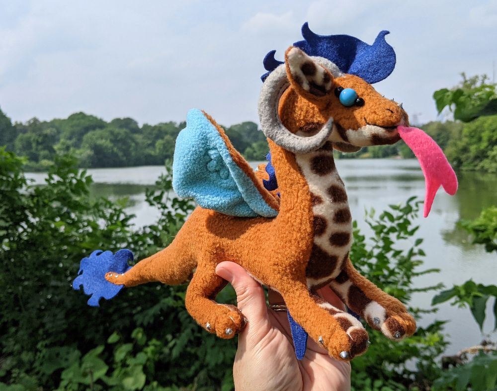 a warm brown dragon with a dark brown/cream patterned underbelly and inner ears, sky blue underwings, dark brown mane/tail/elbow tufts, gray/silver claws/horns/markings, a bright pink tongue, and sky blue eyes