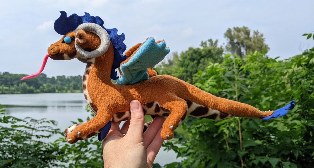 a warm brown dragon with a dark brown/cream patterned underbelly and inner ears, sky blue underwings, dark brown mane/tail/elbow tufts, gray/silver claws/horns/markings, a bright pink tongue, and sky blue eyes
