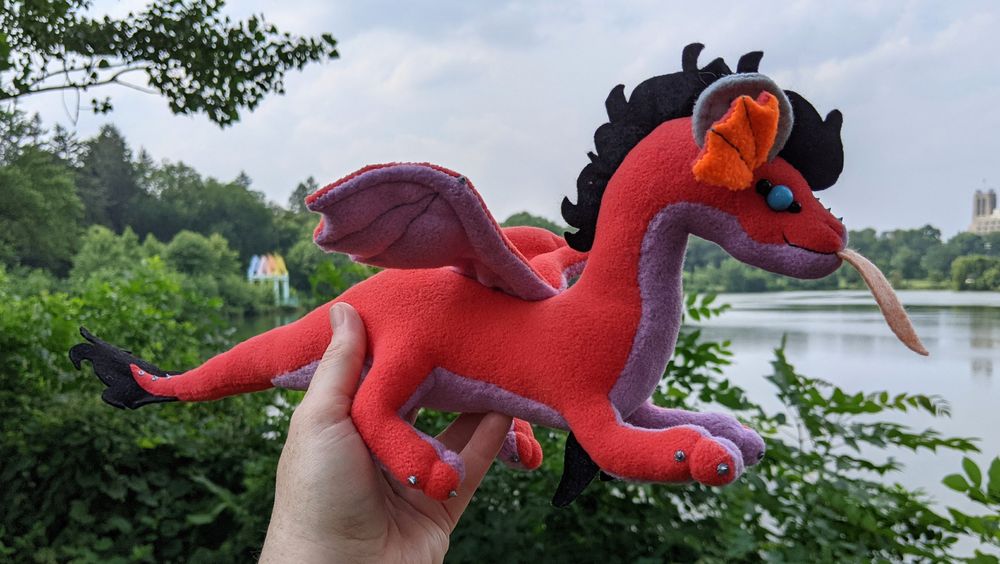 a pink-red dragon plushie with a lilac/light purple underbelly and underwings, bright orange inner ears, grey/silver horns/claws/markings, sky blue eyes, a peach tongue, and a black crest and tailtuft