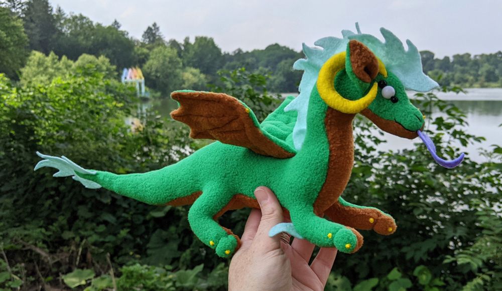 a green dragon plushie with a warm brown underbelly/underwings/inner ears, yellow horns/claws/markings, a purple tongue, aqua mane/tail/elbow tufts, and white eyes