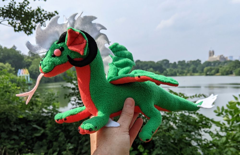 a watermelon colored dragon plushie (bright green with pink-red underbelly & accents, black claws/horns, white mane/tailtip, pink eyes)