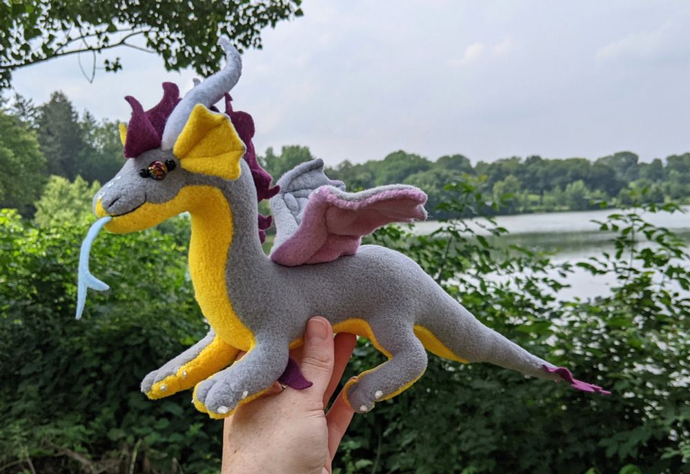 a soft gray dragon plushie with a honey yellow underbelly and inner ears, light purple/lilac underwings, raspberry colored mane/tailtuft/elbow tufts, white horns/claws/markings, and golden purple glass eyes