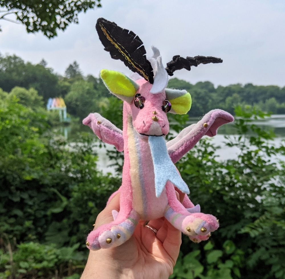a pink dragon with a pastel stripes underbelly, aqua and yellow ears, gray and purple wings, white mane/tail/elbow tufts, gold claws/markings, and black & gold mothlike antennae and golden purple glass eyes